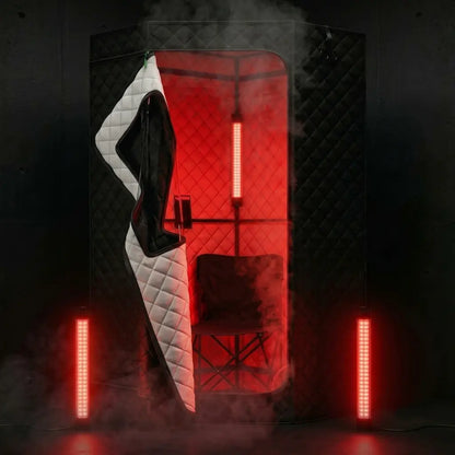 A dark, moody product photo of a privacy sauna cabin with a black quilted exterior, a white quilted side panel, a black interior chair, and glowing red LED light bars on the walls and floor.