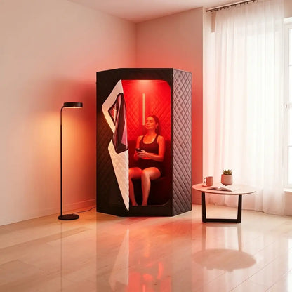 A woman sits inside a black privacy pod sauna in a modern room with warm red and orange lighting. The room has beige walls, a floor lamp, and a small side table with a cup and plant.
